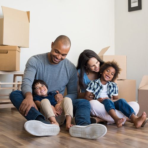 Happy family with two children having fun at new home. Young multiethnic parents with two sons in their new house with cardboard boxes. Smiling little boys sitting on floor with mother and dad.